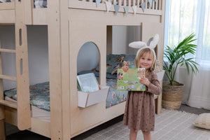 Child standing in a room with a wooden bunk bed and a plant.