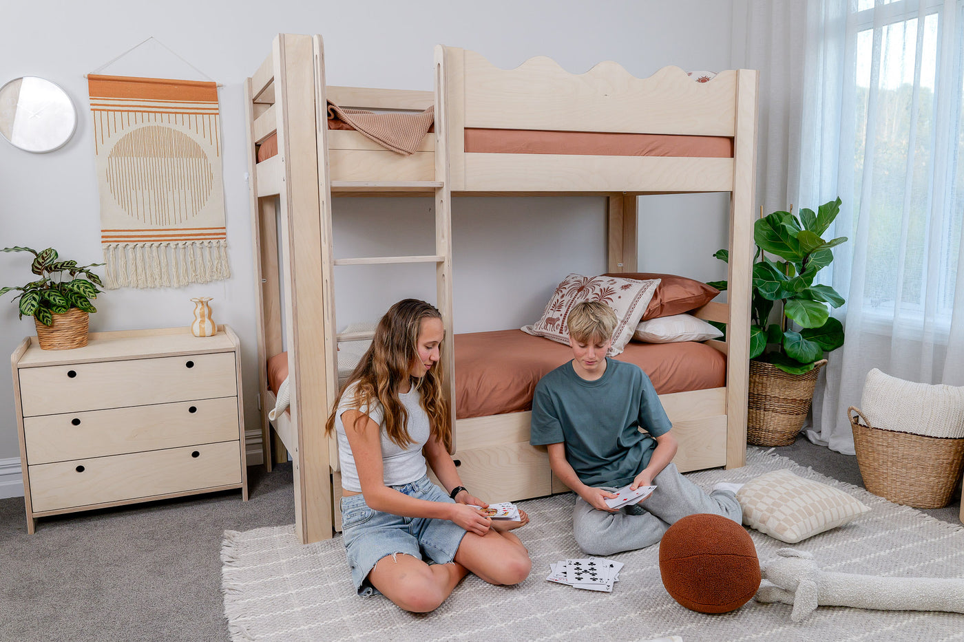 Two children are sitting on the floor next to a set of Wavy bunk beds in a room with plants and furniture.